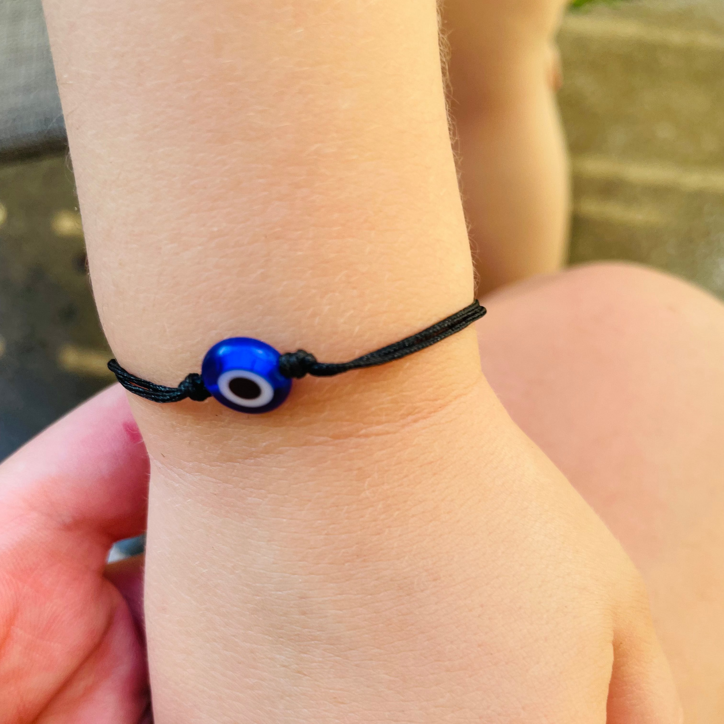 Toddler hand wearing a thread bracelet with an evil eye charm, held by another hand.