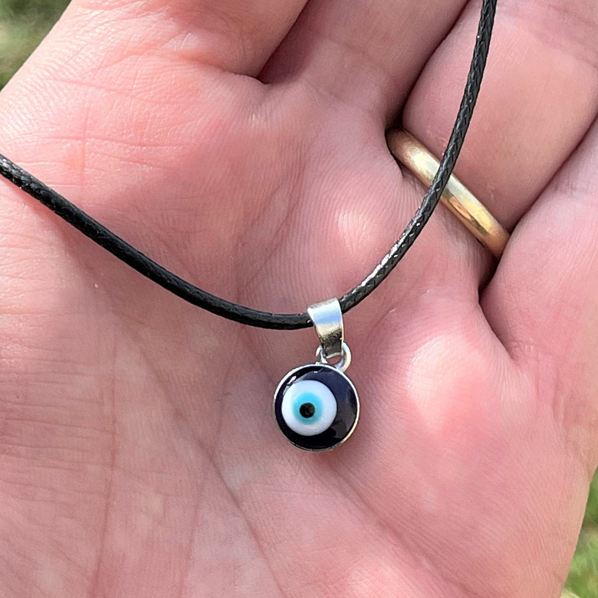 Hand holding a necklace with a tiny round eye pendant on a black cord against a grassy background