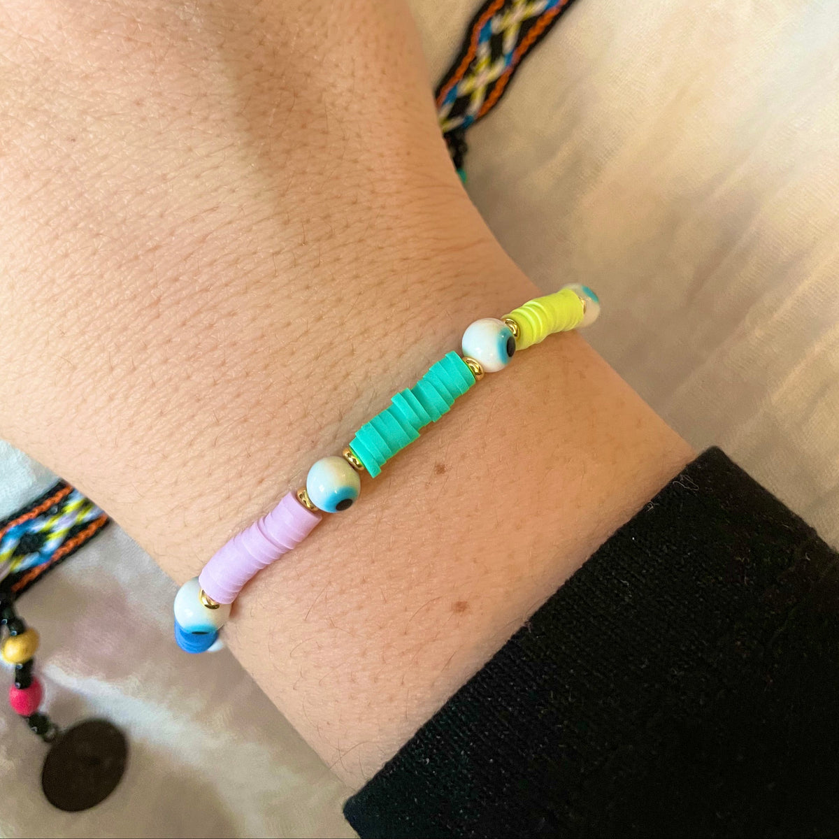 Colorful beaded bracelet with white evil eye beads and neon yellow, green and violet heishi beads on a wrist with a blurred background