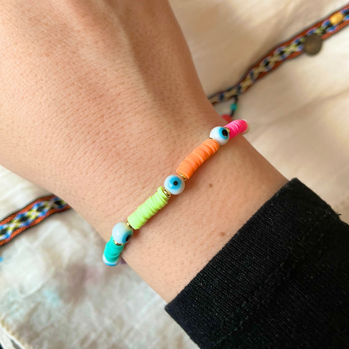Colorful beaded bracelet with white evil eye beads and pink, orange, yellow and green heishi beads on a wrist with a blurred background