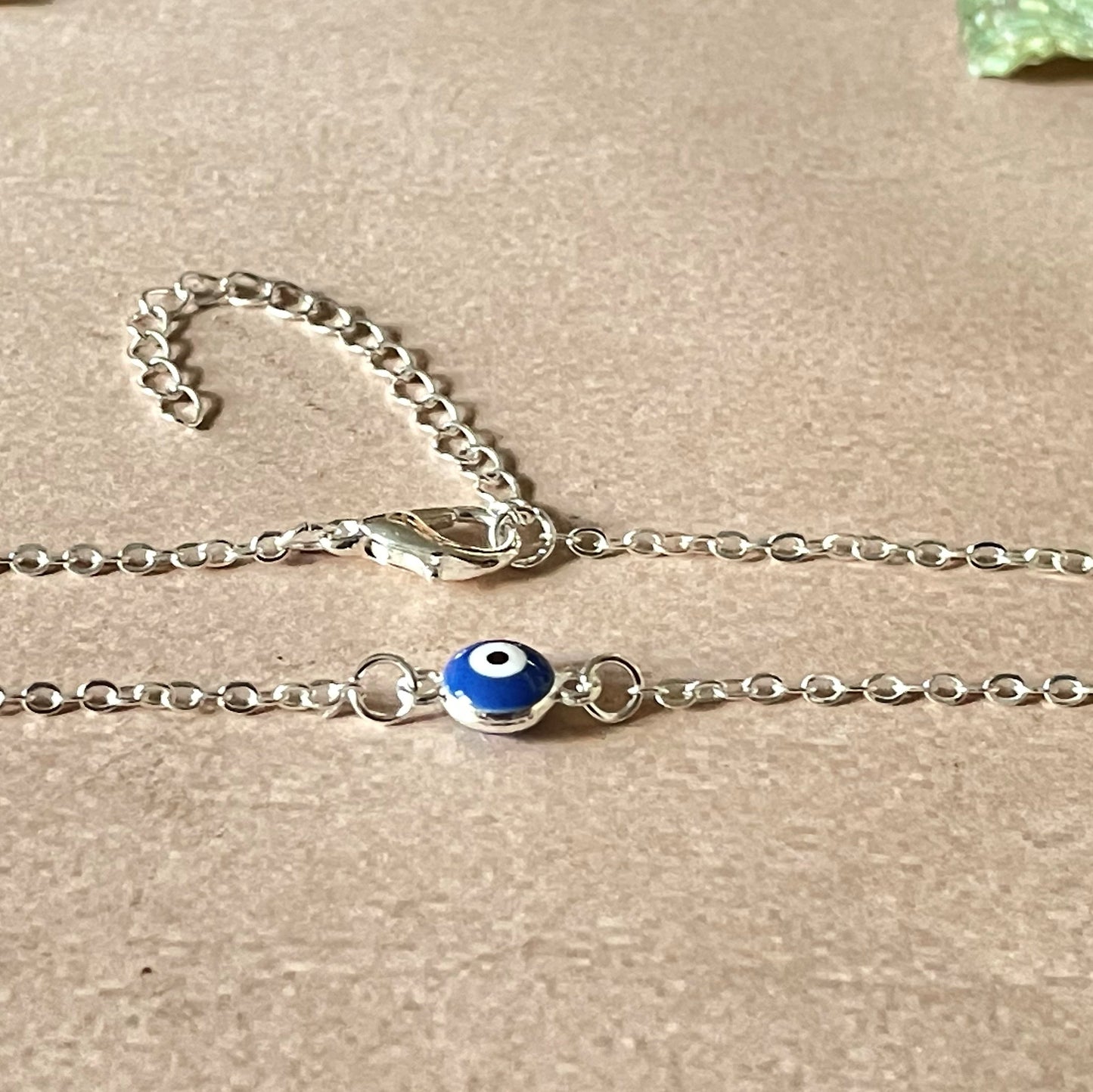 Close-up view of silver chain anklet with a blue evil eye charm on a beige background