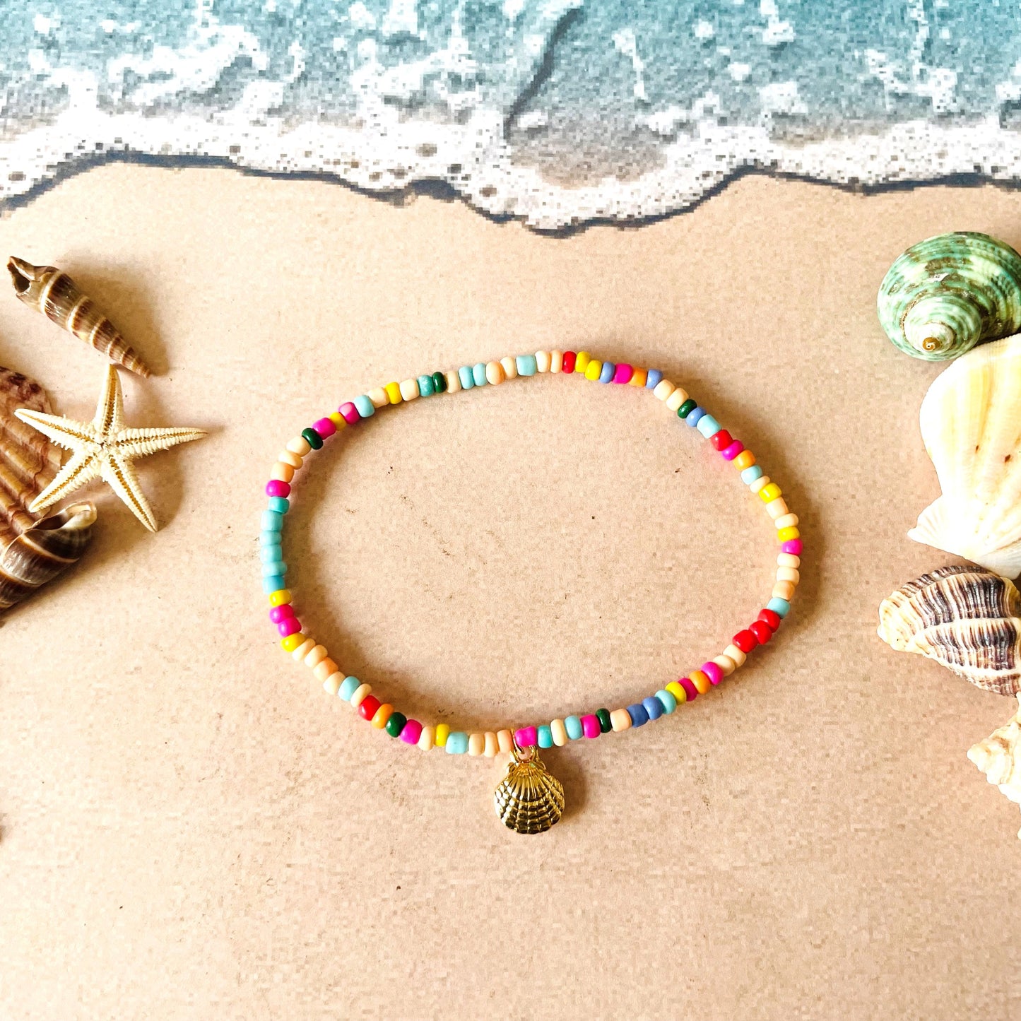 Rainbow seed bead anklet with a gold shell charm on sand with seashells and starfish