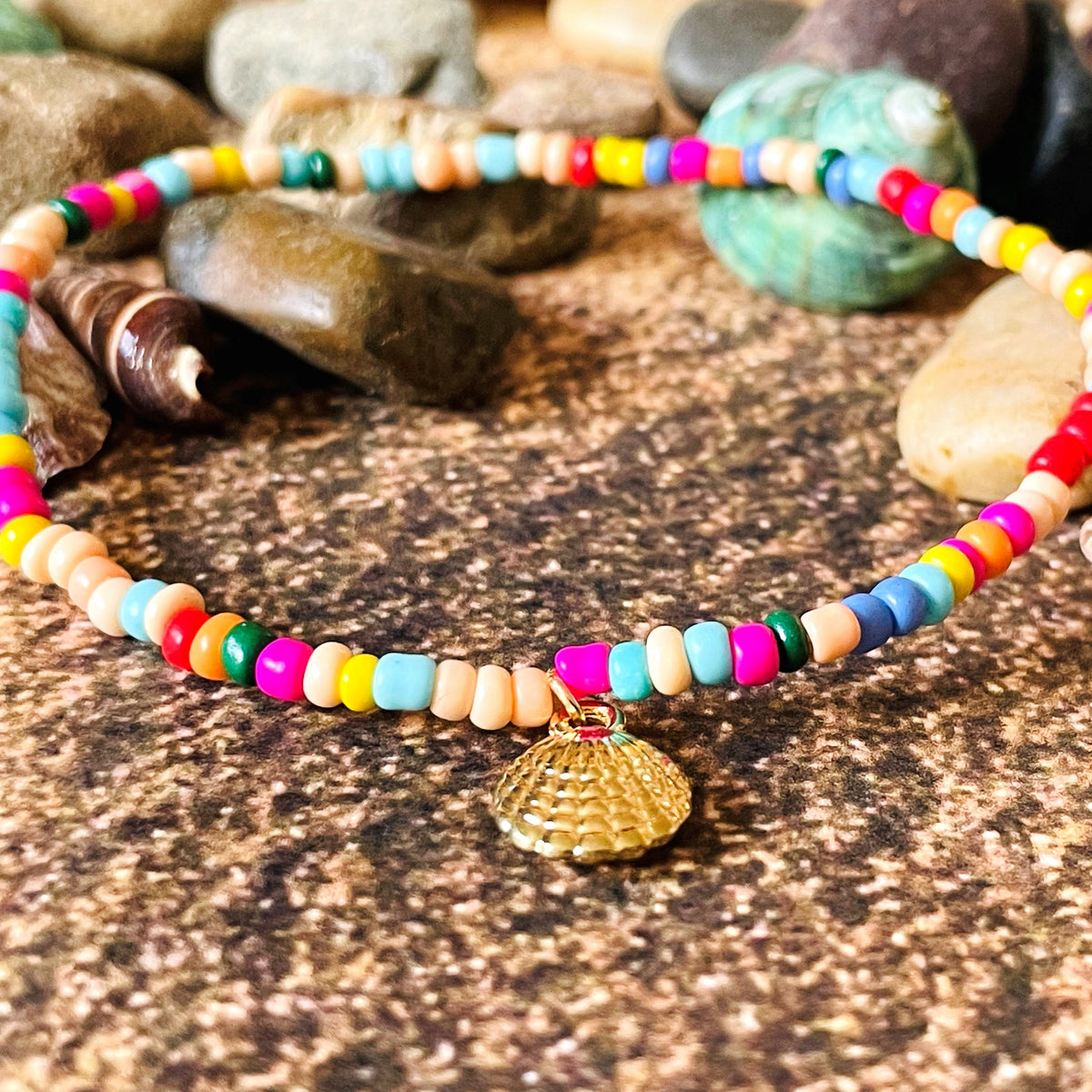 Colorful beaded stretch anklet with a gold shell charm on a textured surface with stones and shells.