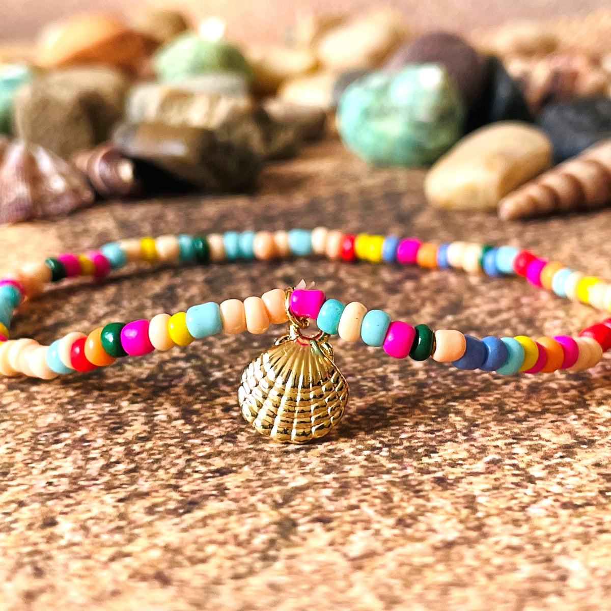 Front view of multicolored beaded anklet with a gold charm on a textured surface with shells in the background