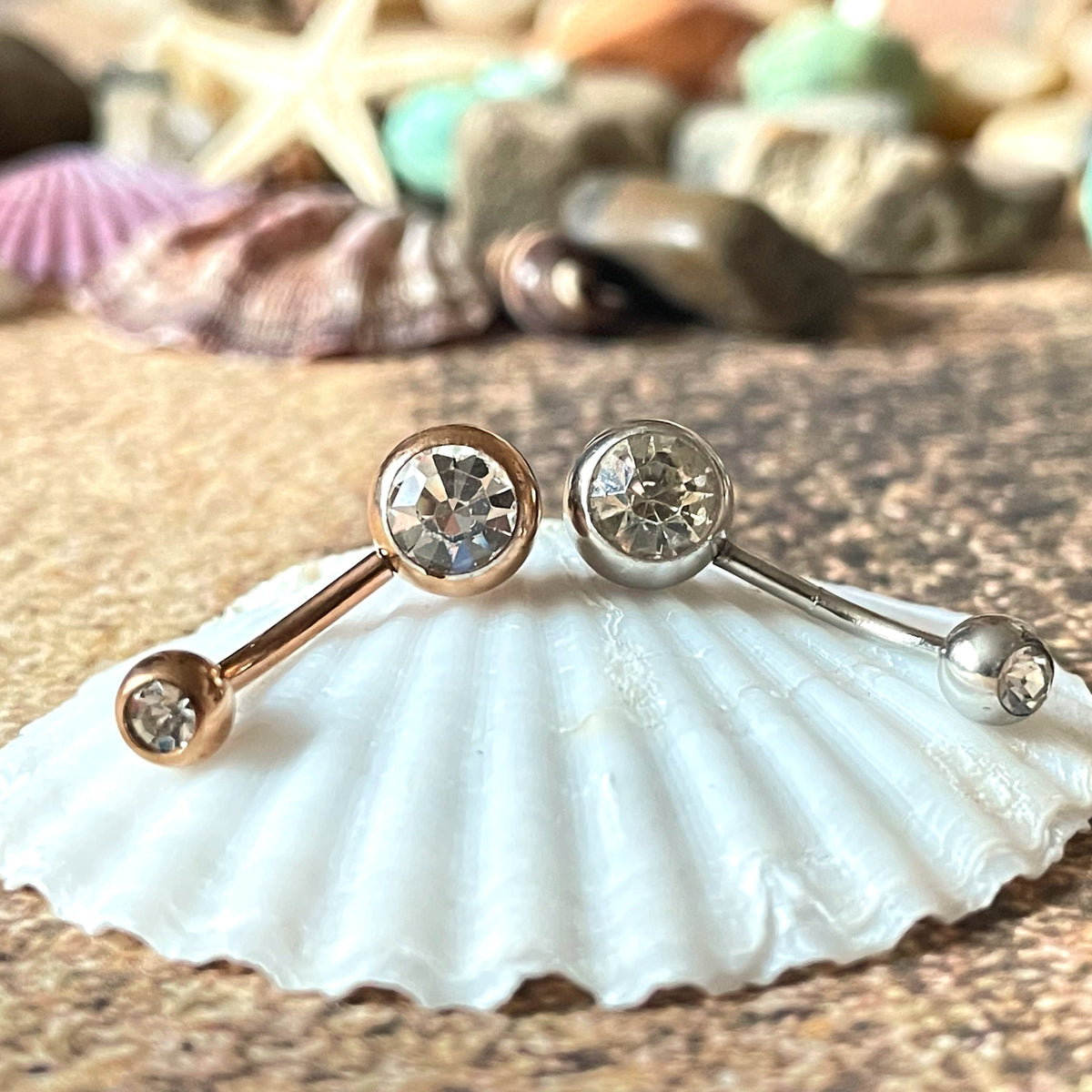 Pair of belly rings, one silver and one rose gold, on a shell with a beach background
