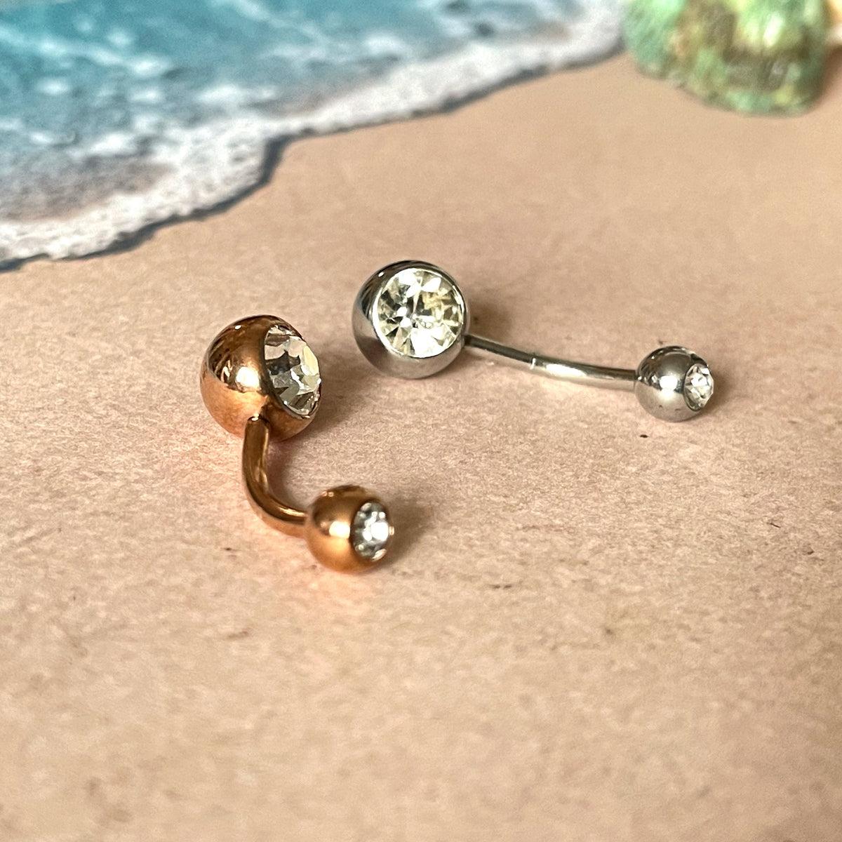 Gold and silver belly button rings on sand with ocean waves in the background