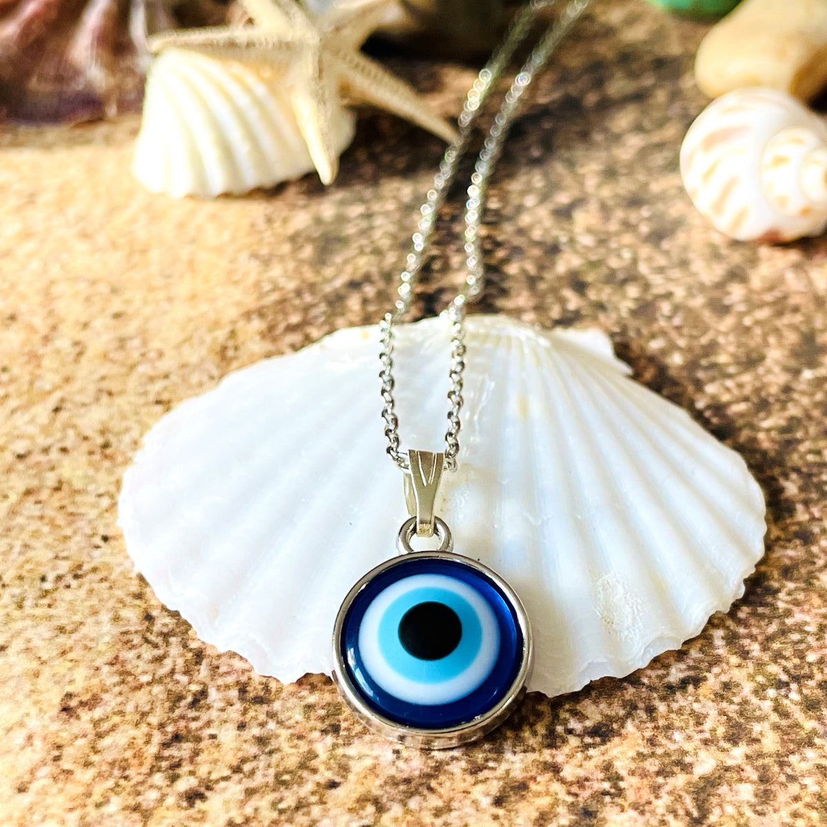 Silver necklace with a blue eye pendant on a seashell against a sandy background