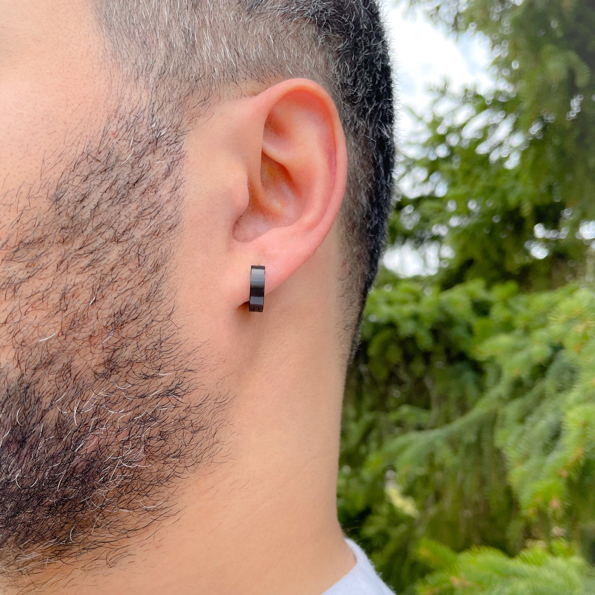 Man wearing black hoop earrings with a smooth high-polish finish with a blurred green outdoor background