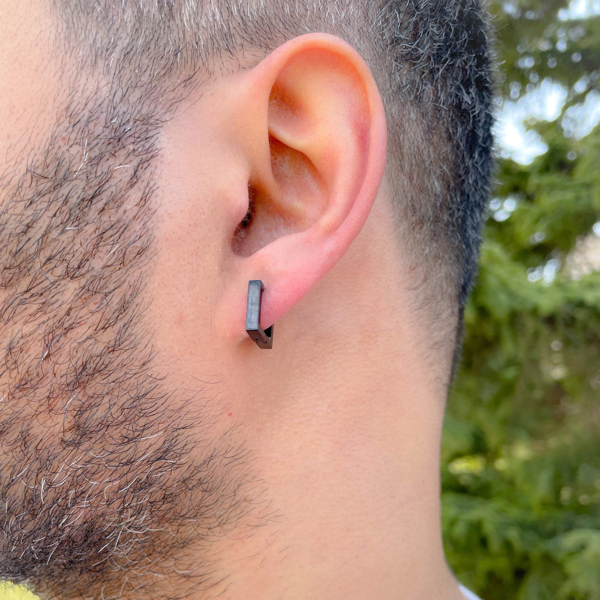 Person wearing black square hoop earrings with a blurred green outdoor background