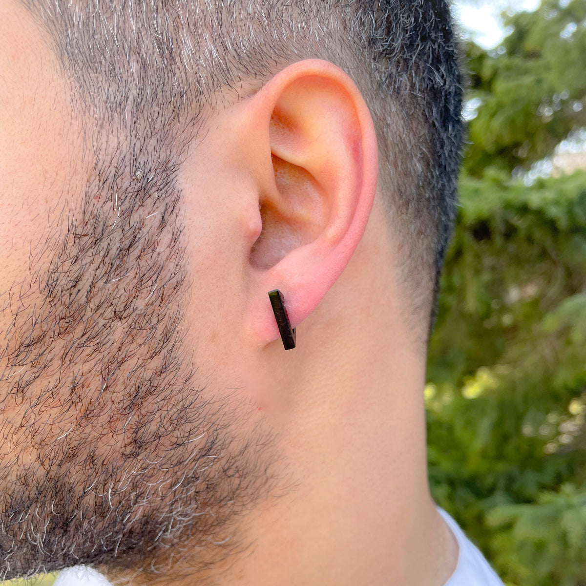 Close-up of a person wearing a black triangle hoop earring outdoors with greenery in the background