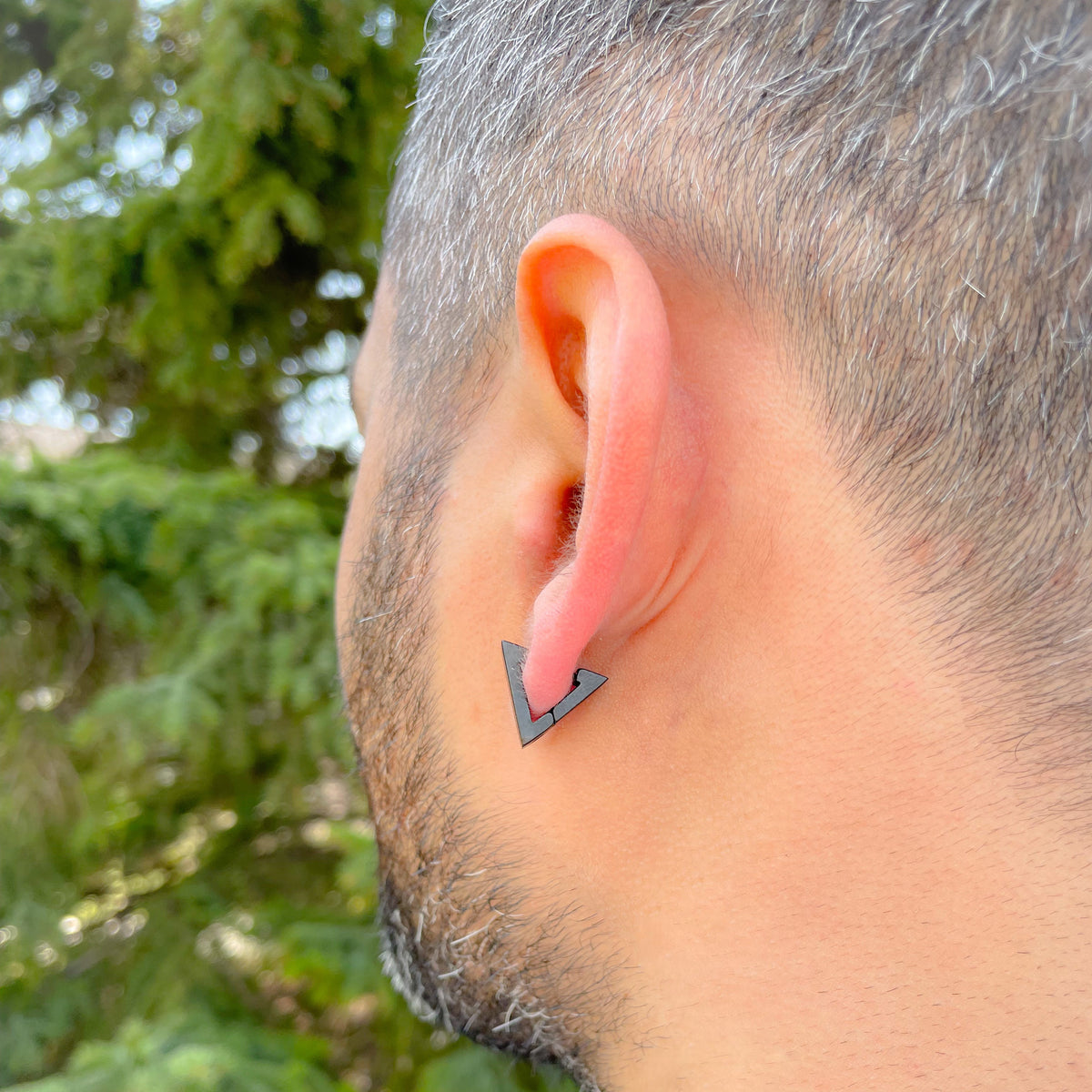 Person wearing geometric black triangle hoop earrings with a blurred green outdoor background