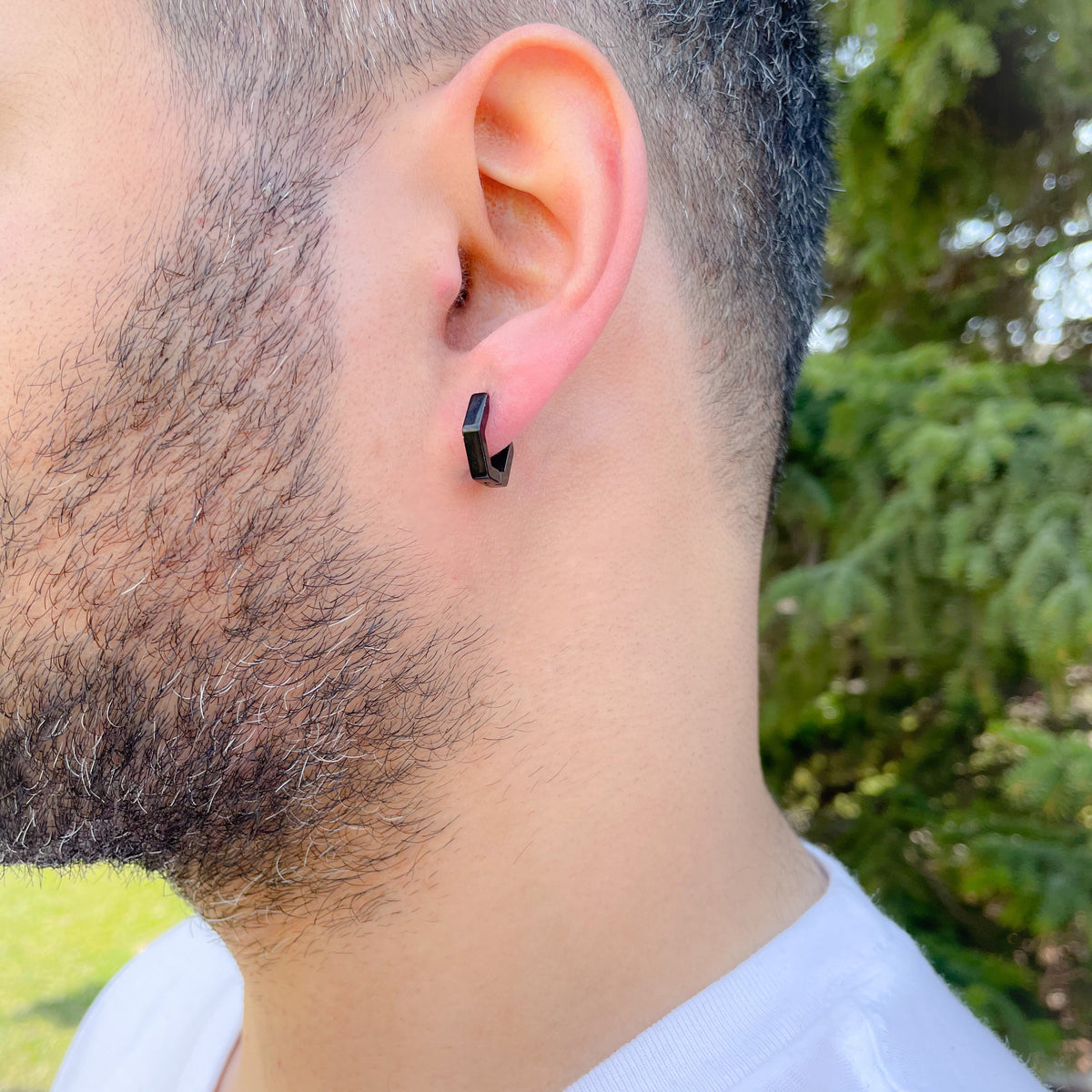 Person wearing a black hexagon shaped hoop earring with a blurred green outdoor background