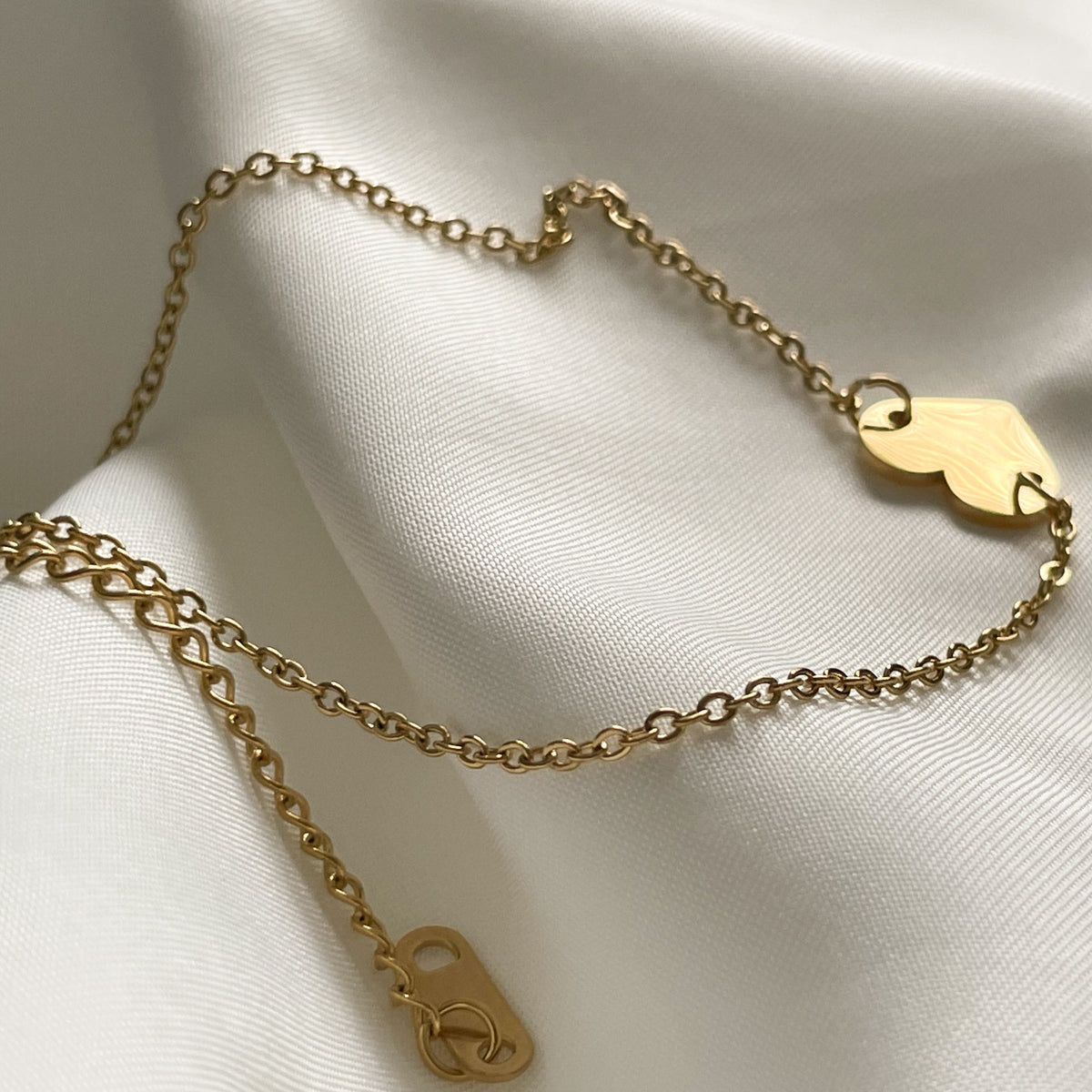 Gold chain ankle bracelet with heart charm and extender chain on a white fabric background