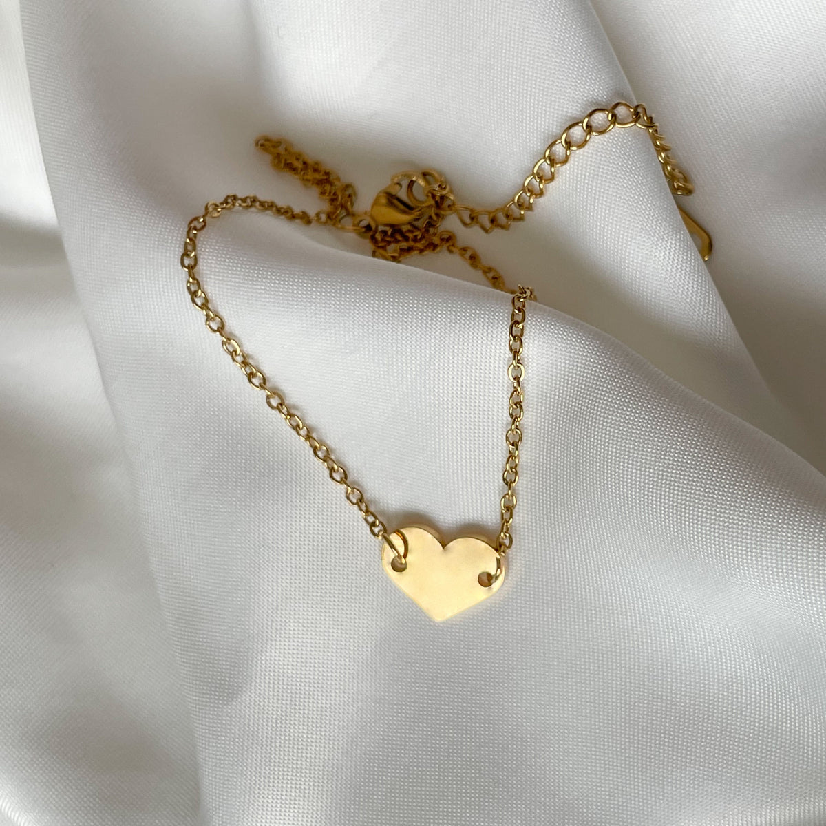 Gold flat heart-shaped anklet with a delicate cable chain on a white fabric backgroundh