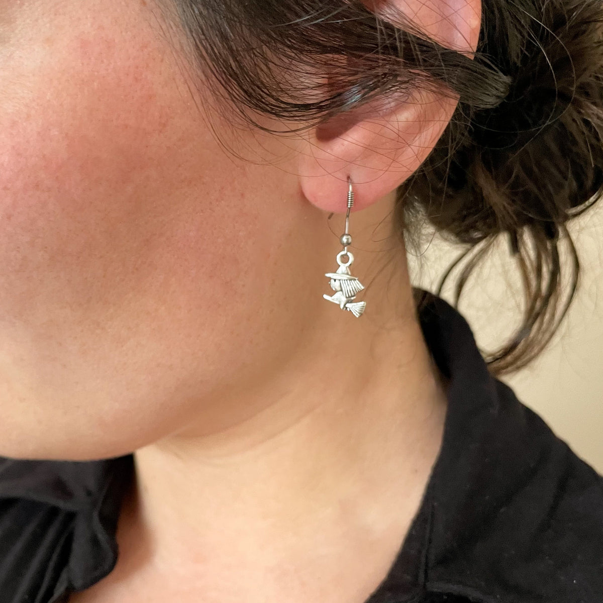 Close-up of a person wearing cute silver earrings with witch on a broom charm with a neutral background