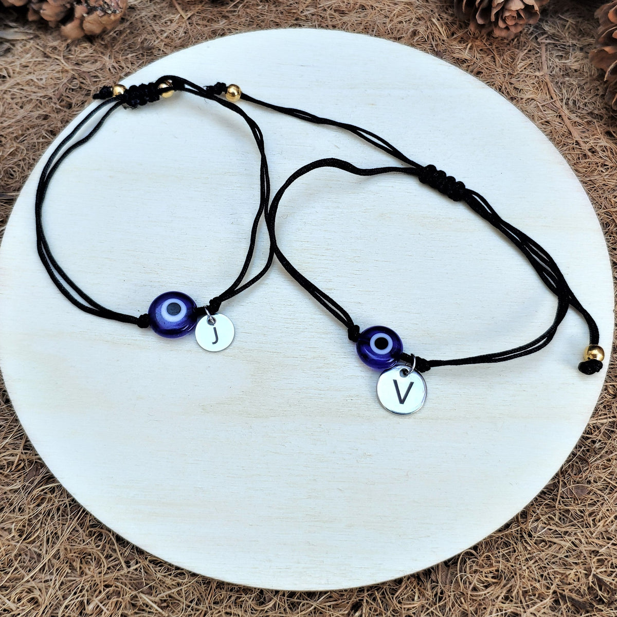 Two black evil eye bracelets with letter charms on a white stone surface, surrounded by pine cones and white flowers.