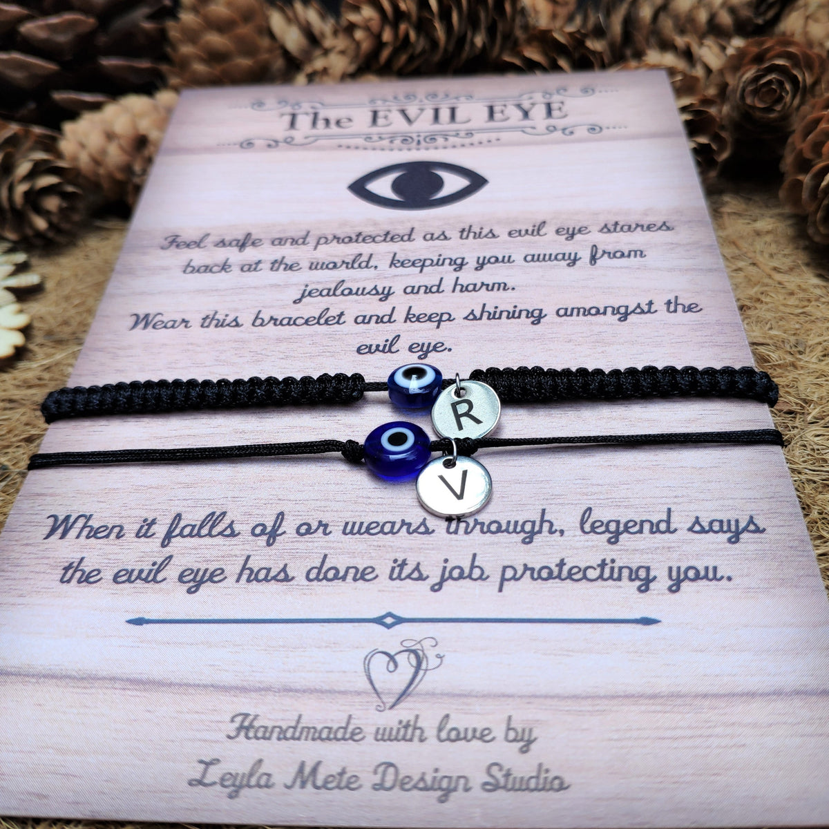 Two bracelets, one braided and one thread with evil eye charm and silver initial disc on a card with text about the evil eye, surrounded by pine cones.