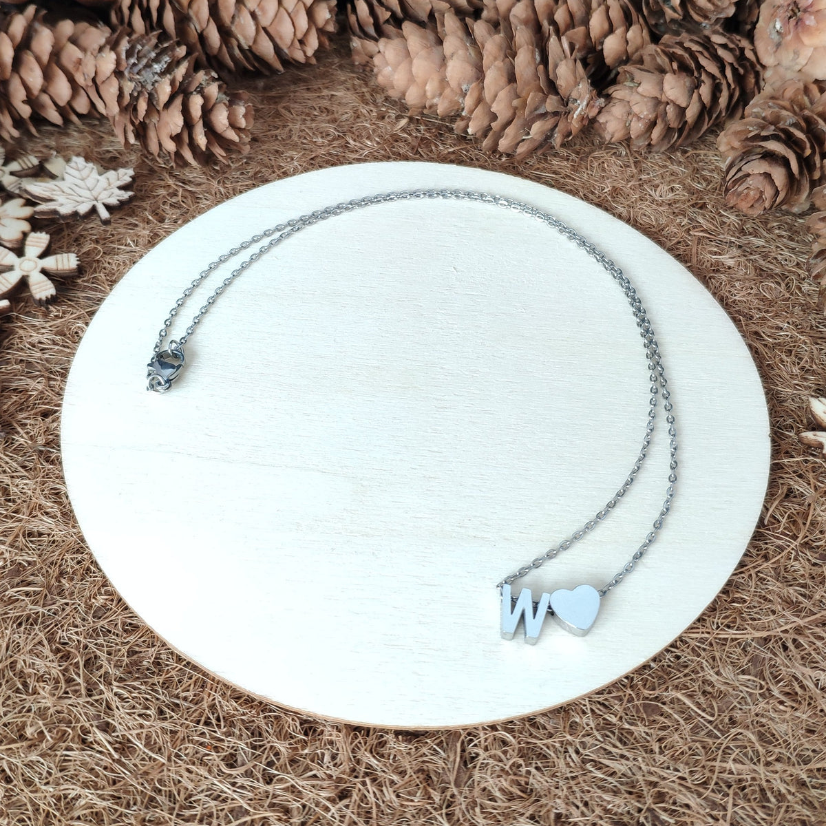 Necklace with heart and letter 'W' pendant on a white plate surrounded by pine cones and snowflakes.
