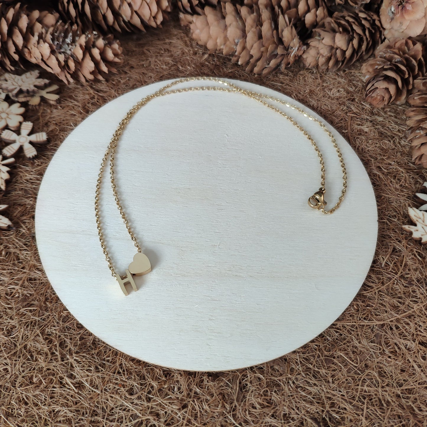 gold necklace with a heart and letter 'H' on a wooden white plate surrounded with pinecones