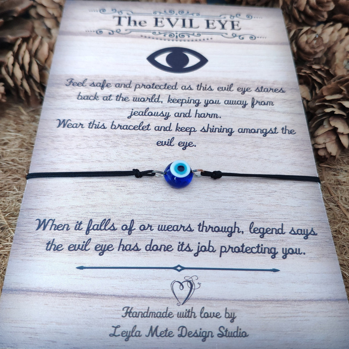 Round evil eye bracelet on a brown card with text about its legend and branding surrounded by pine cones.