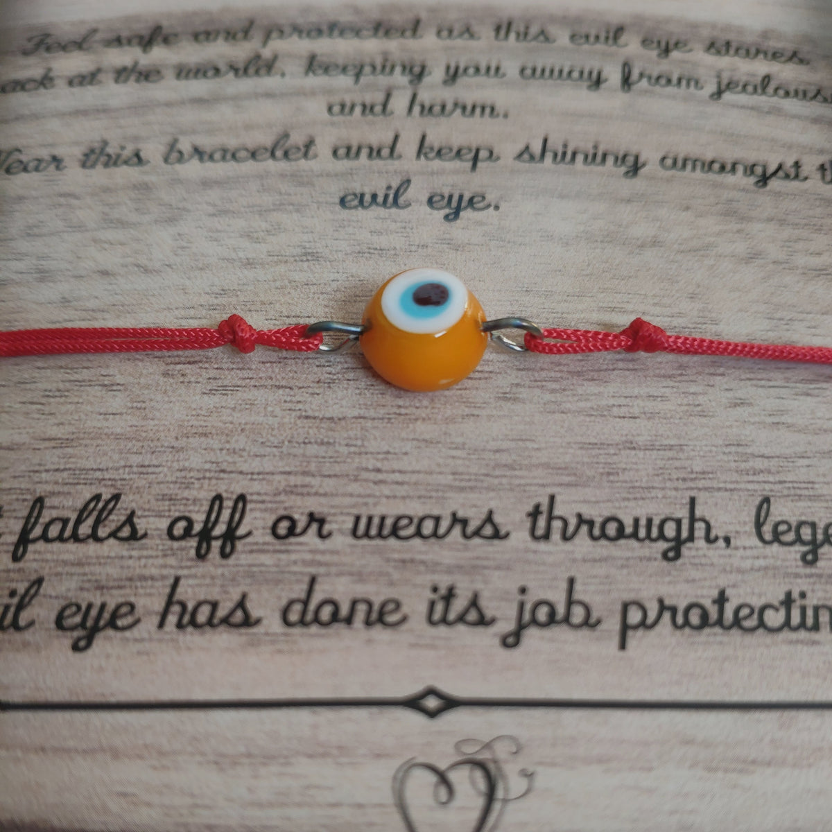 Red String bracelet with orange evil eye charm on a brown card with text