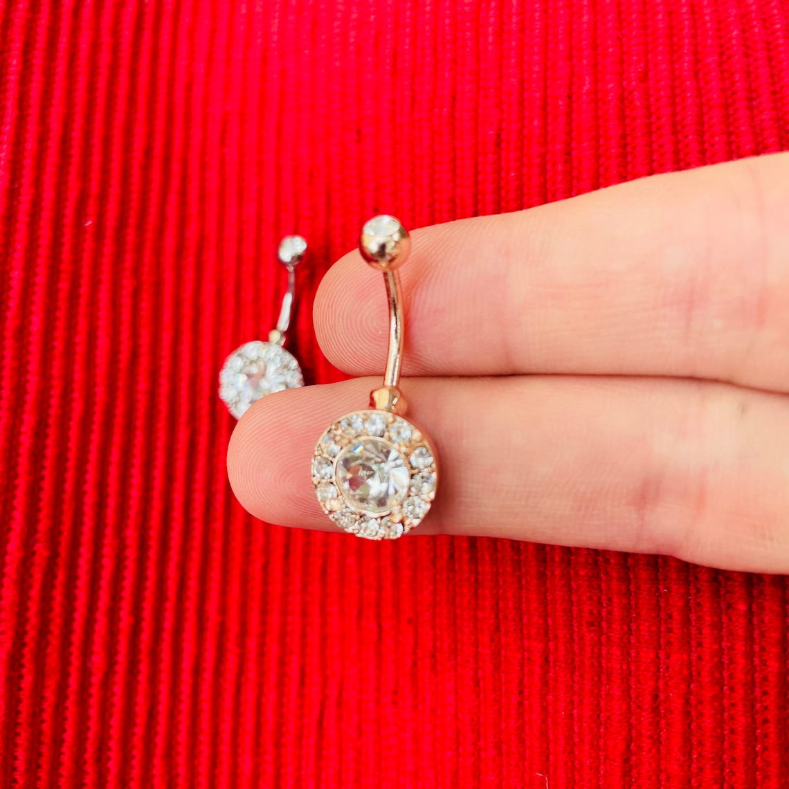 Rose gold crystal belly ring held on hand, against a red background