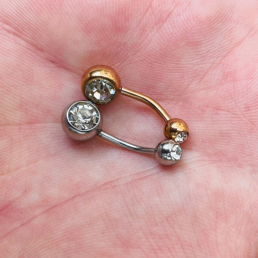 One silver and one rose gold belly button rings with two clear gemstones on a hand
