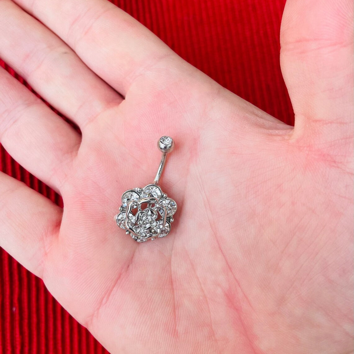Silver belly button ring with rose-shaped design and embedded crystals on a hand against a red background