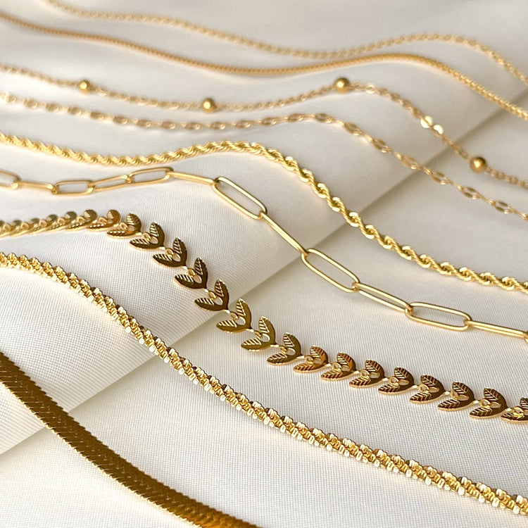 Angled close up of 9 different chain necklaces made of 18k gold PVD plated stainless steel, displayed on white satin. From left to right:  3 mm herringbone, sparkle rope, chevron, paperclip, 2 mm rope, mirror (sequin), satellite, box and cable.
