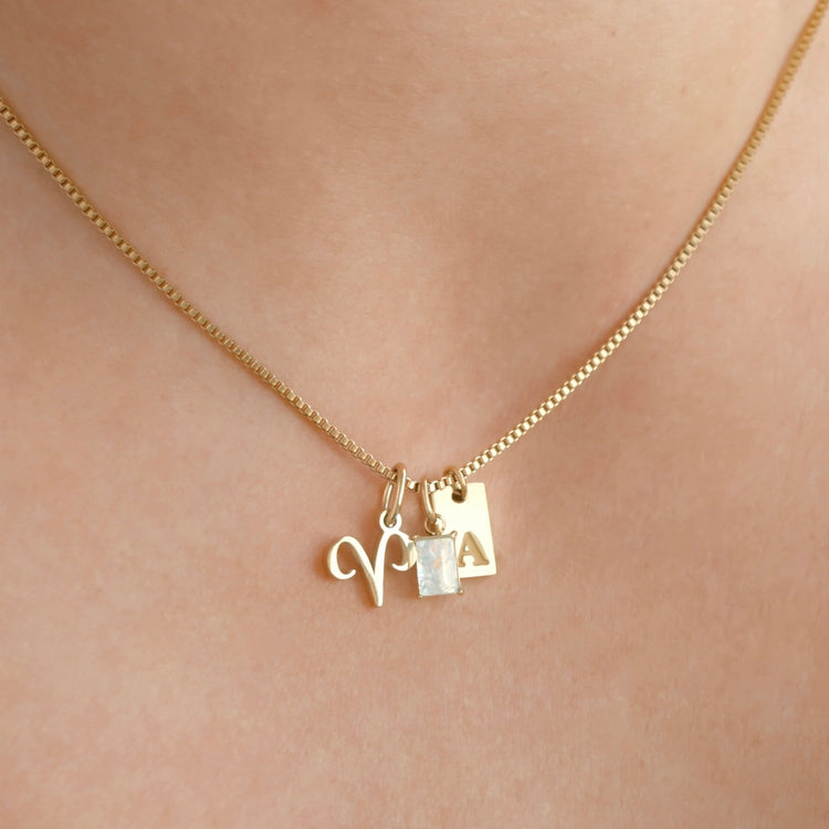 Dainty gold necklace charms, handcrafted in sterling silver in a beige ring holder section of a jewelry box. The charms are arranged in 3 rows, a smiley face, angel, heart, padlock, sun, diamond crescent, butterfly, north star and moon tag charm.
