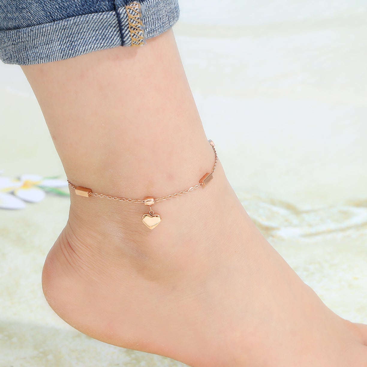 Rose gold anklet with a heart charm and rectangular beads on a foot with a soft background