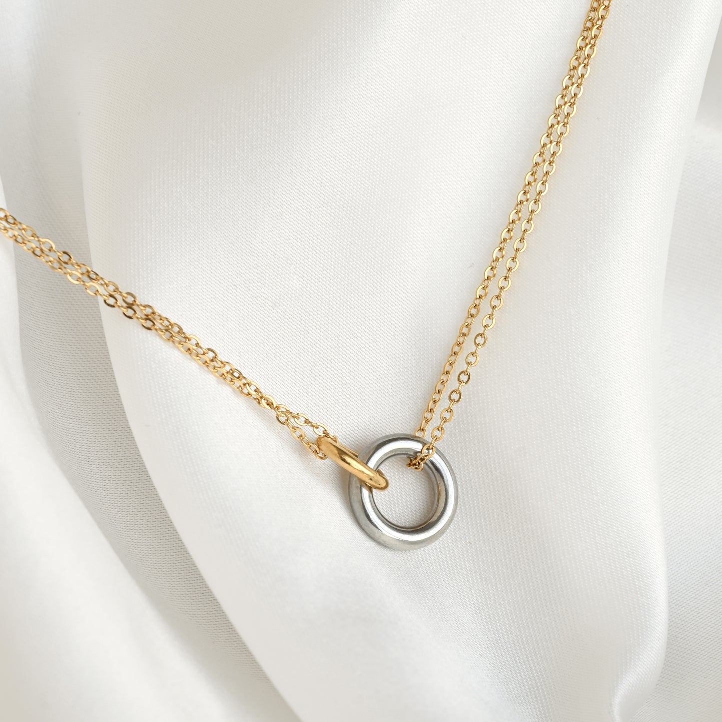 A delicate mixed metal chain necklace with a double interlocking ring pendant, featuring one gold ring and one silver-toned ring, displayed on a soft white satin fabric.
