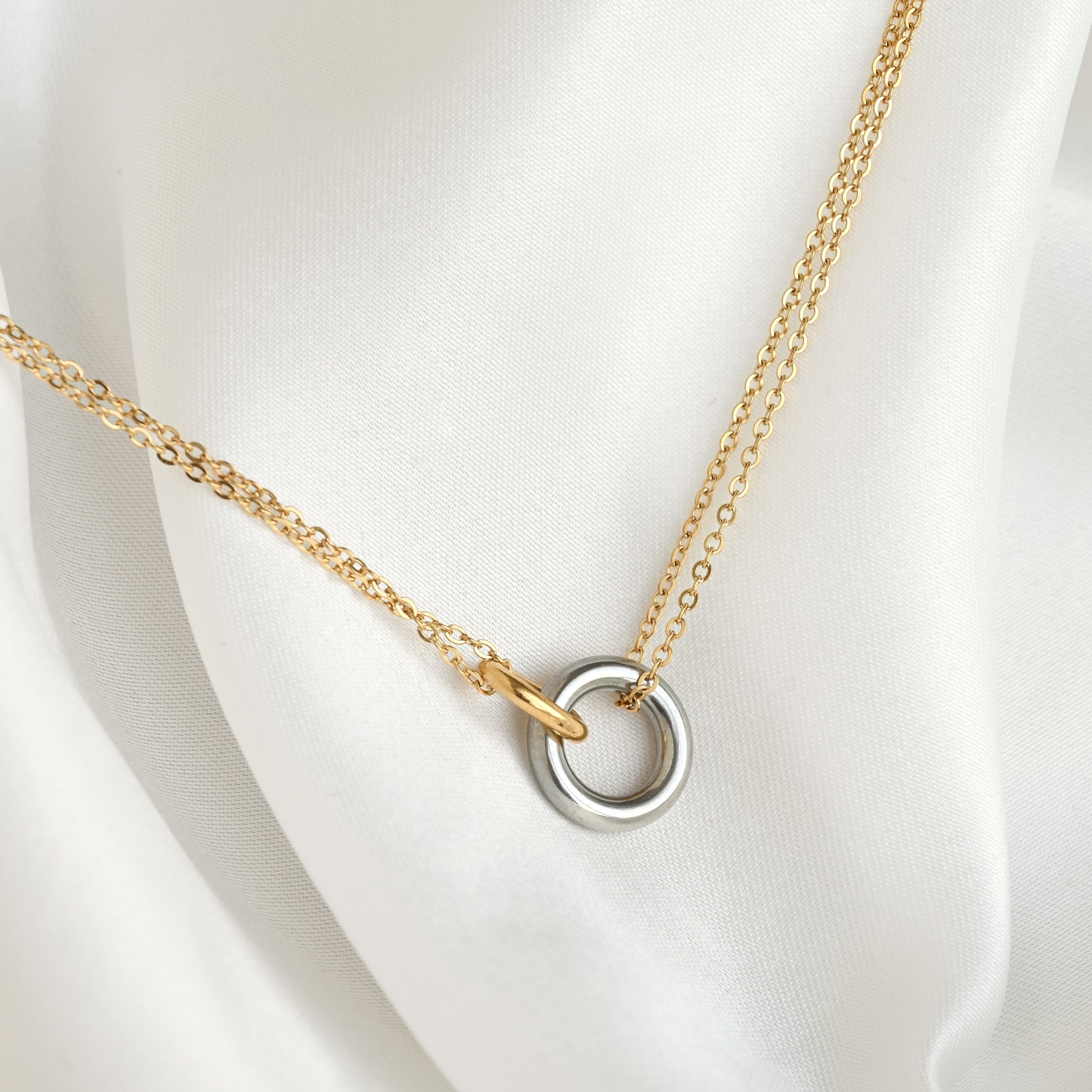 A delicate mixed metal chain necklace with a double interlocking ring pendant, featuring one gold ring and one silver-toned ring, displayed on a soft white satin fabric.