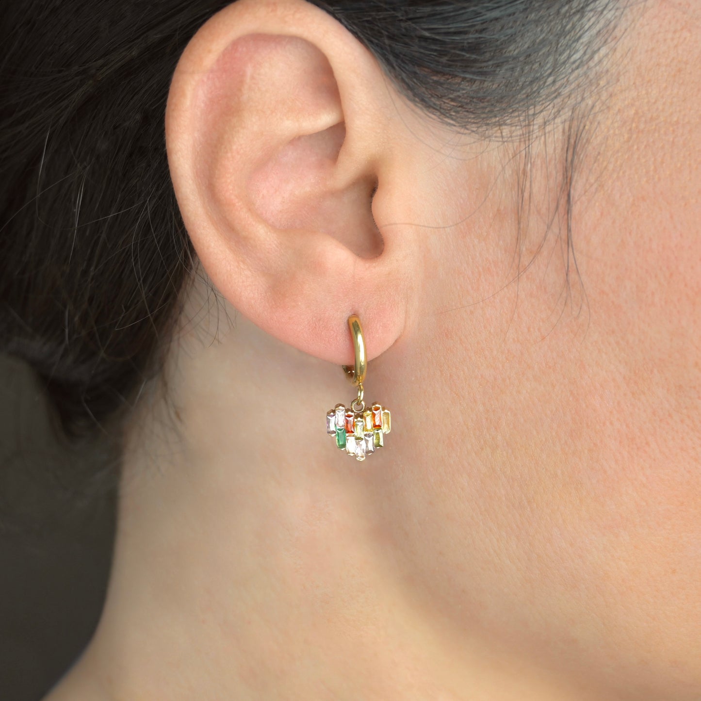 Close-up of an ear wearing a gold hoop earring with a heart charm adorned with colorful  rectangular stones.