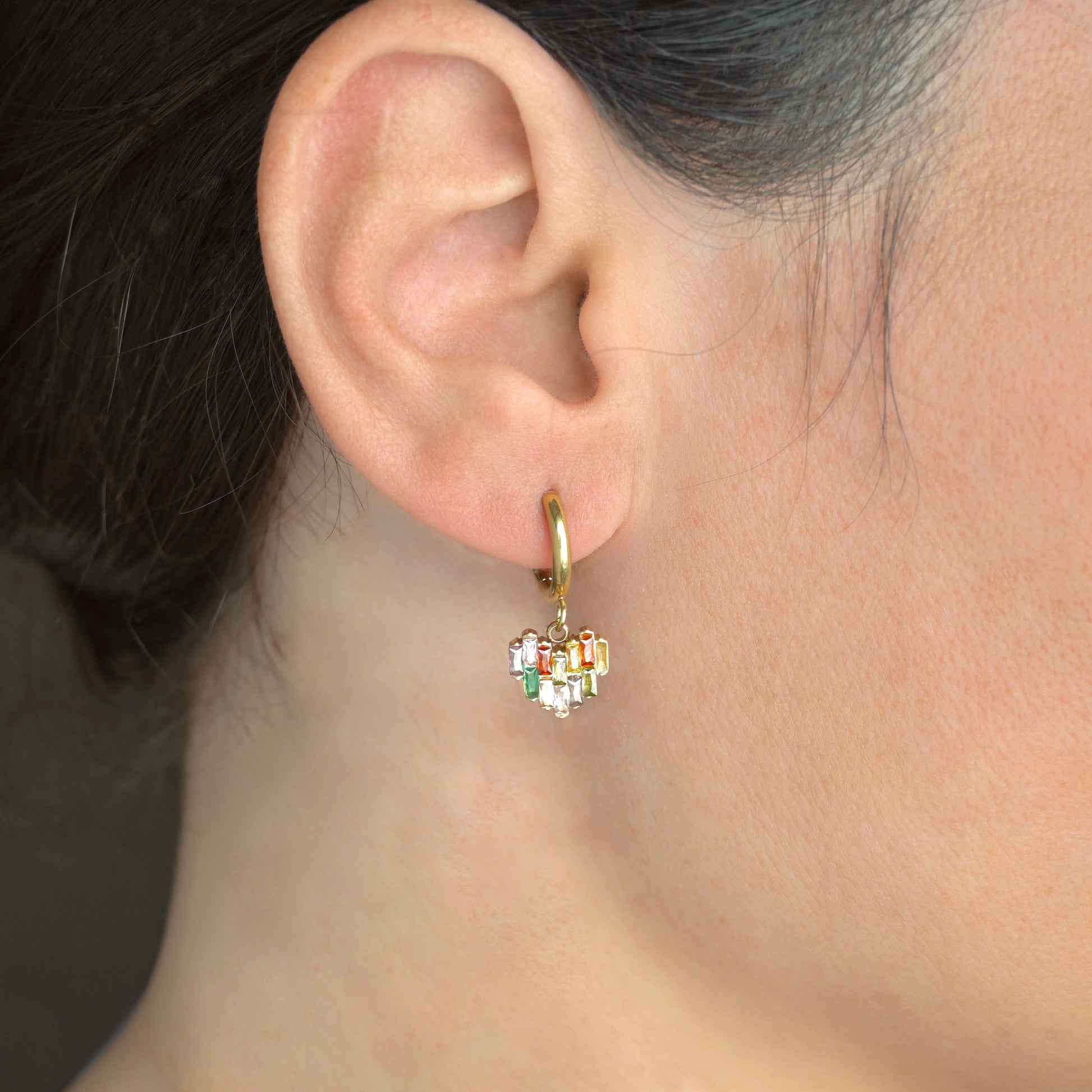 Close-up of an ear wearing a gold hoop earring with a heart charm adorned with colorful  rectangular stones.