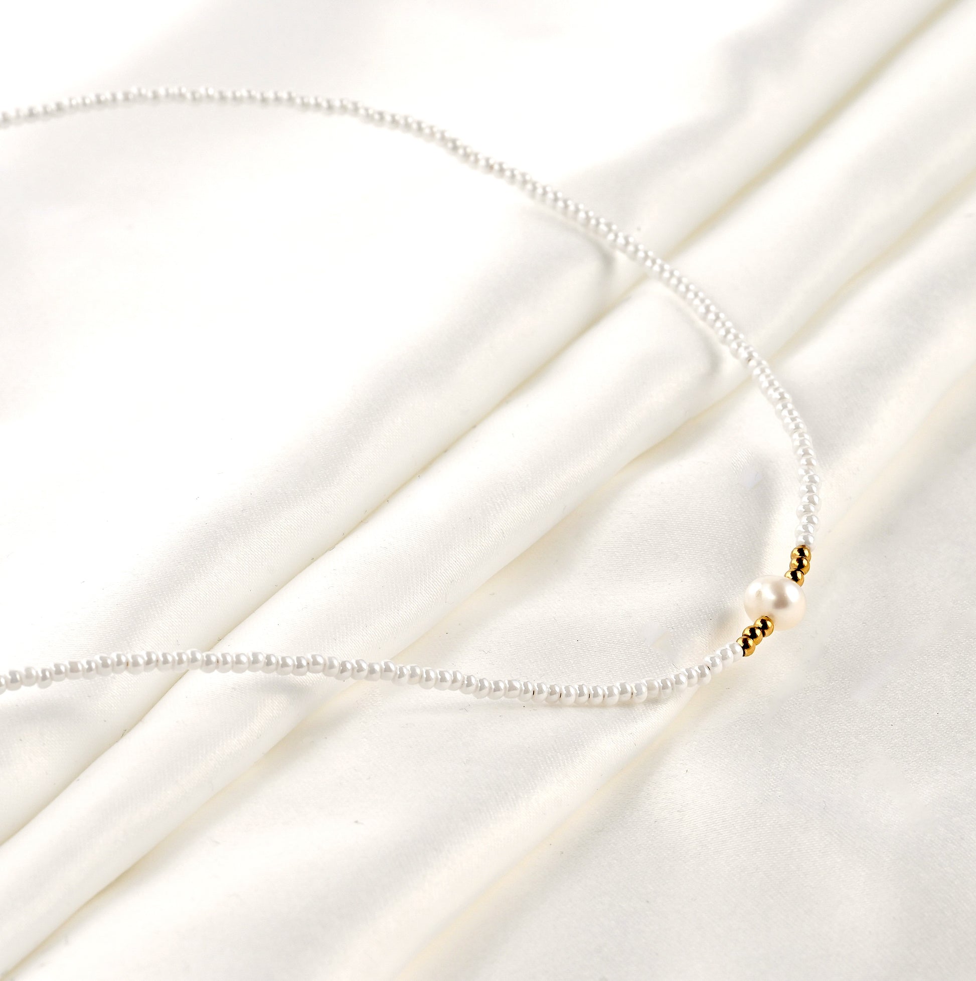Delicate white seed bead necklace with a natural pearl centerpiece and 3 gold beads on each side on a white fabric backgrounf
