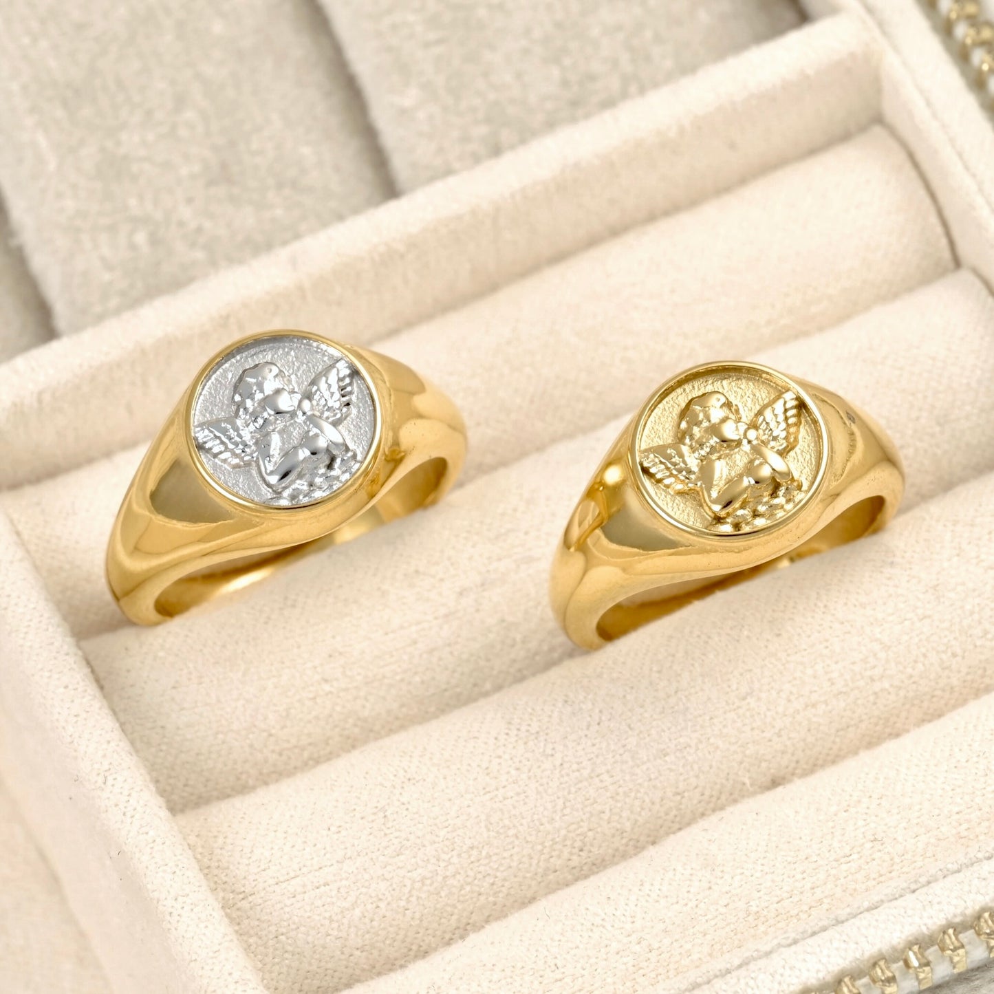 Two signet rings displayed in a cream-colored jewelry box. One ring is yellow gold with a silver-toned winged emblem on its oval face, and the other is yellow gold with a gold-toned winged emblem.