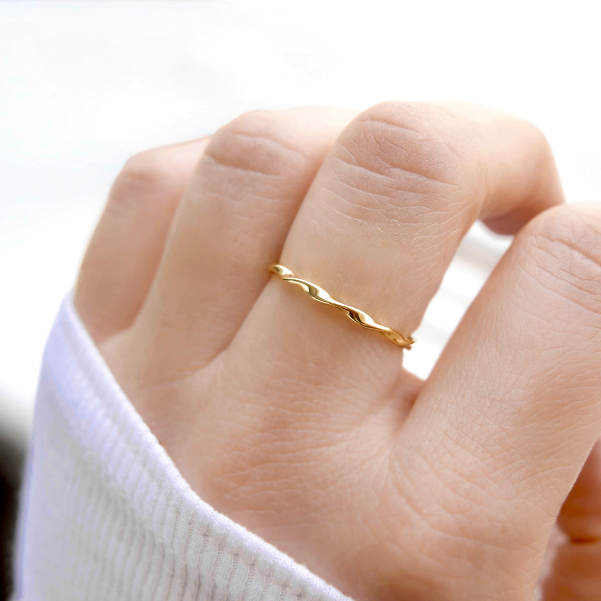 A close-up of a hand wearing a thin, twisted gold band ring, with a white ribbed sleeve visible at the wrist.