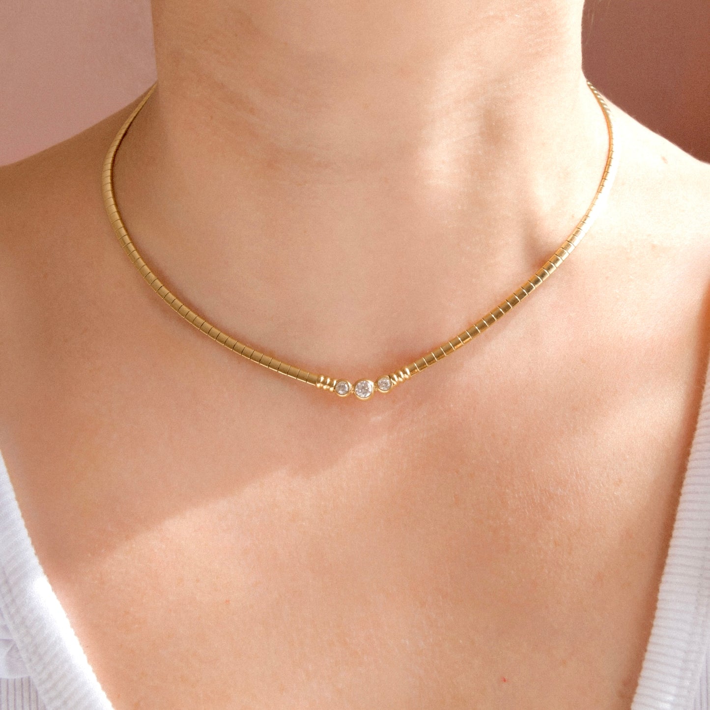 A close-up of a person's neck and upper chest wearing a sleek gold chain necklace with a small clear gemstone at the center, against a soft pink background.