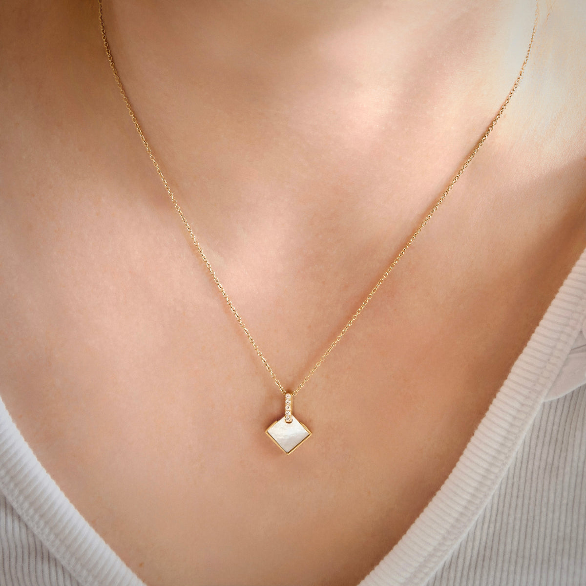 A delicate gold chain necklace with a small, polished diamond-shaped pendant with mother of pearl inlay, worn on a mode and a bar with cz stones holding the square, on a woman's neck over a white ribbed top.