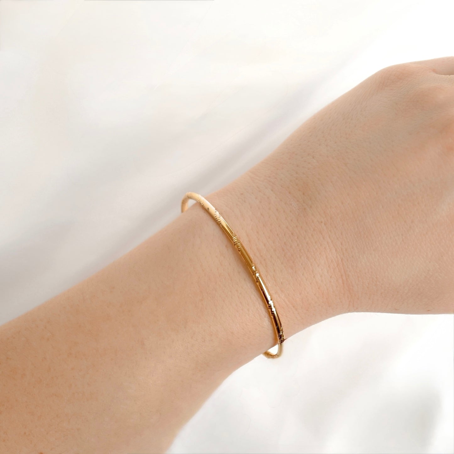 A thin gold bangle bracelet with a textured, diagonal line engraved finish worn on a person's wrist against a soft white background.