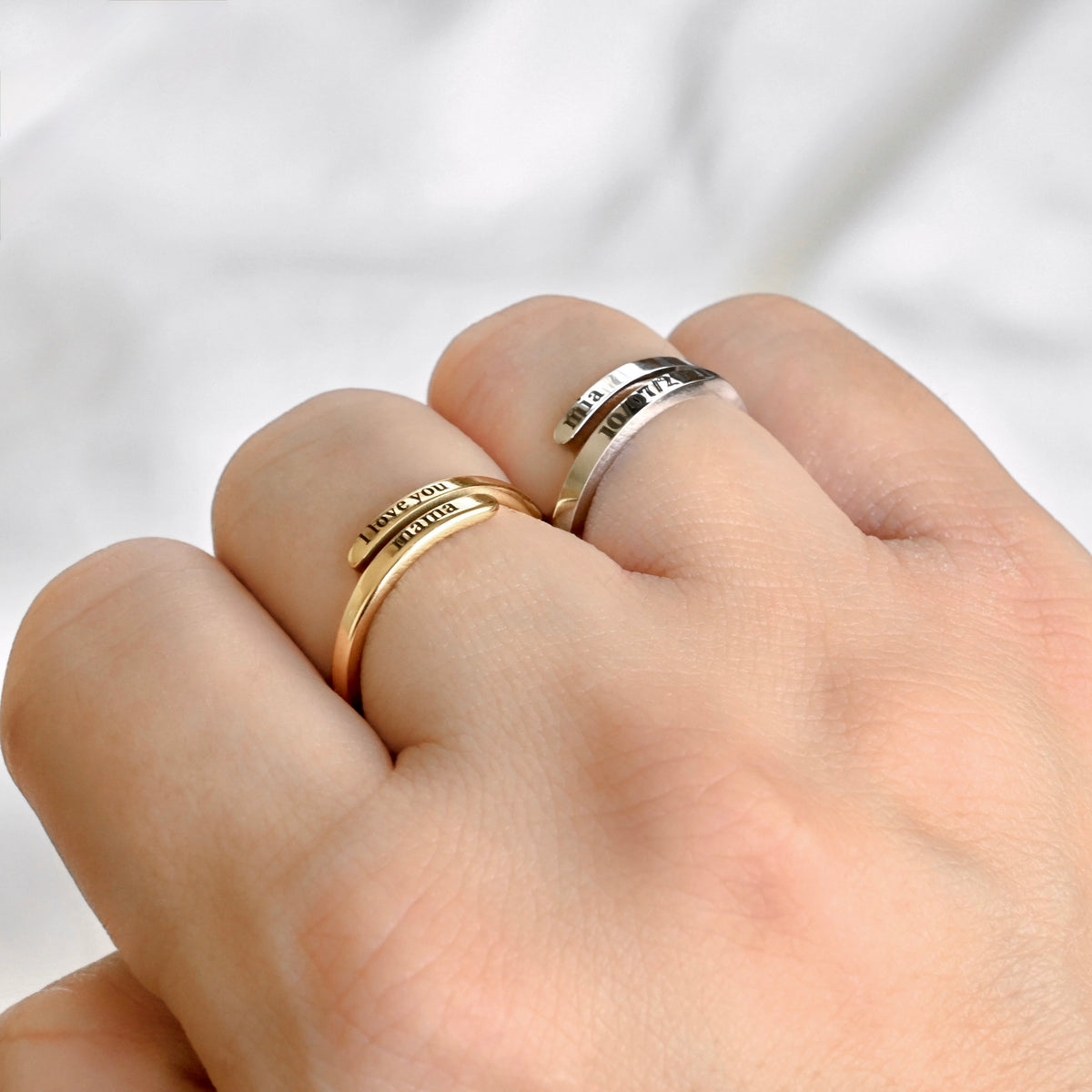 A close-up of a hand wearing two slim wrap-style rings: one gold-toned ring engraved with 'I LOVE YOU' and 'mama,' and one silver-toned ring engraved with 'mia' and a date.