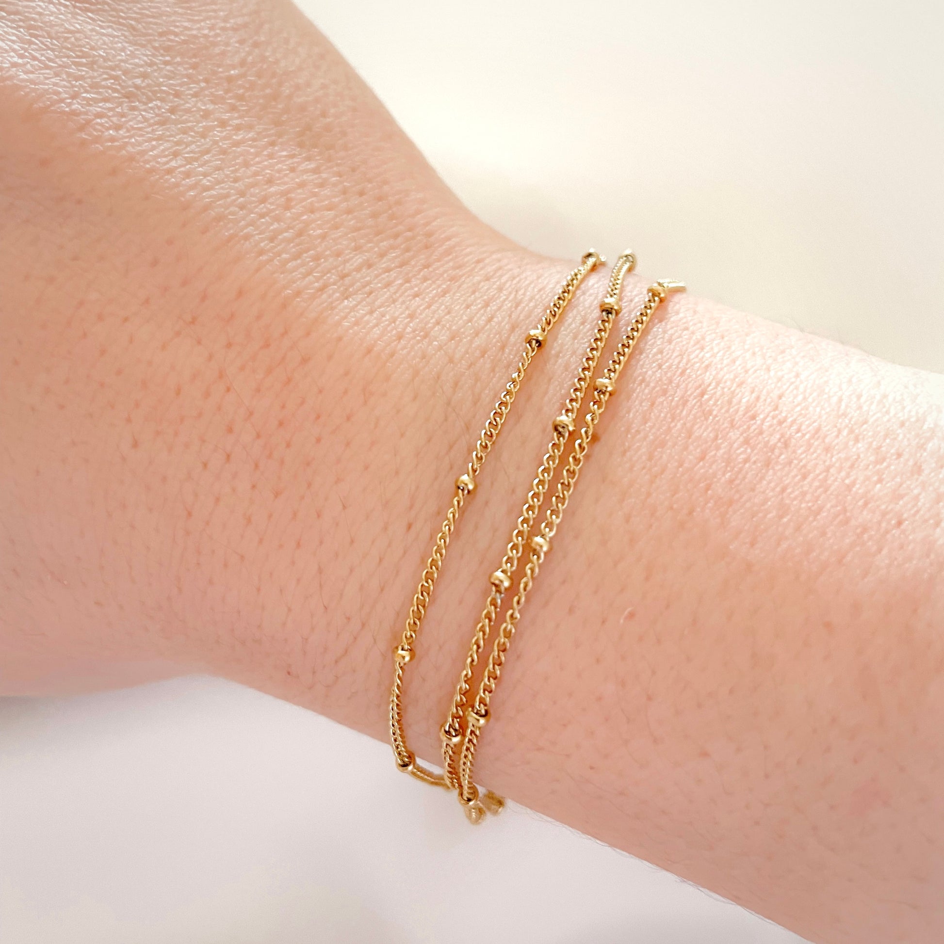 Three-layered dainty gold beaded bracelet on a wrist against a neutral background