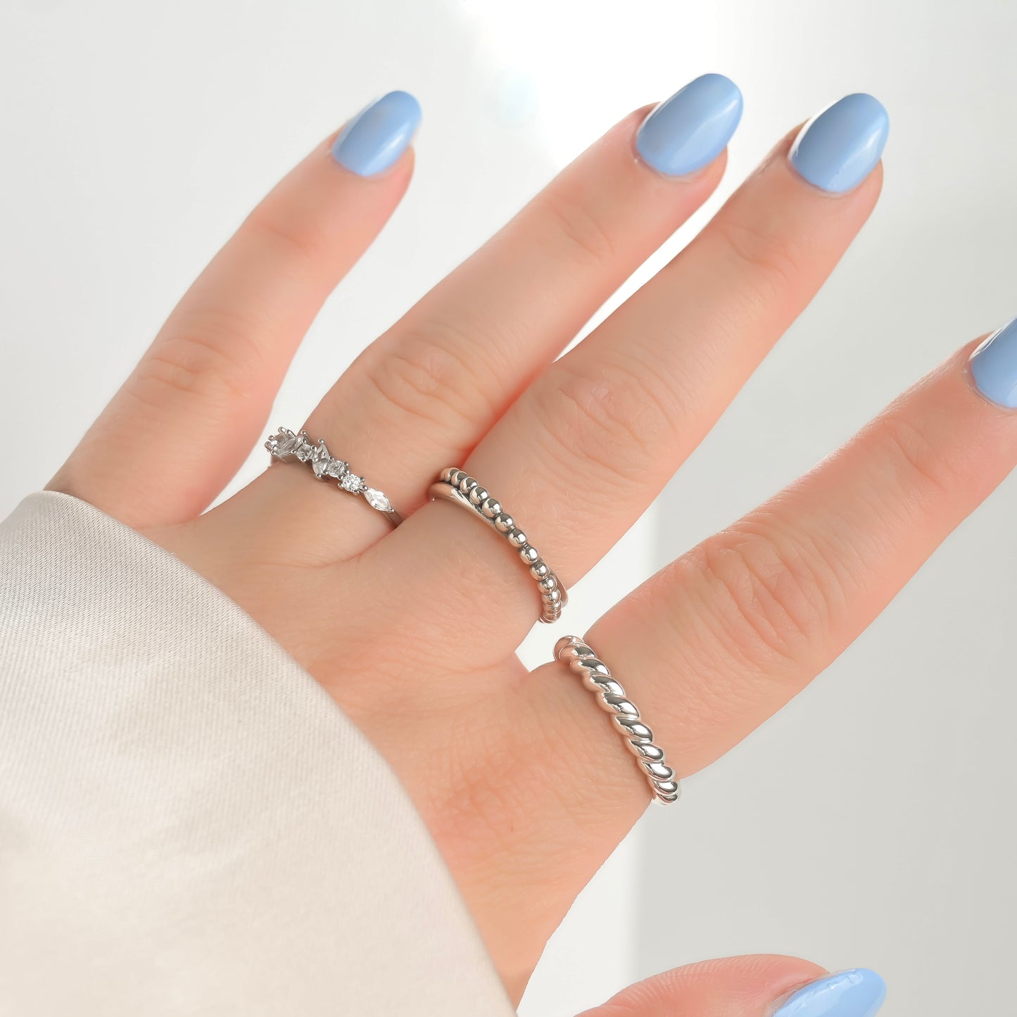 Hand wearing three silver rings with light blue nail polish on a white background