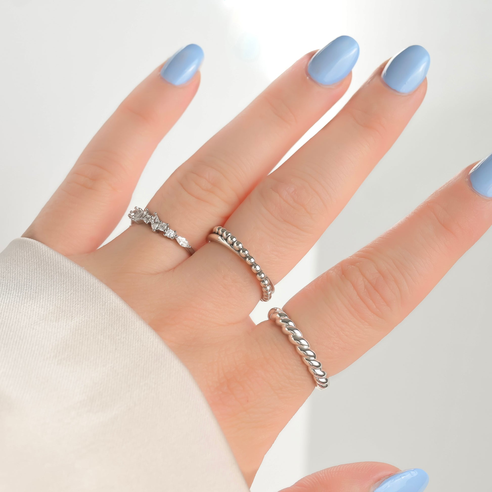 Hand wearing three silver rings with light blue nail polish on a white background