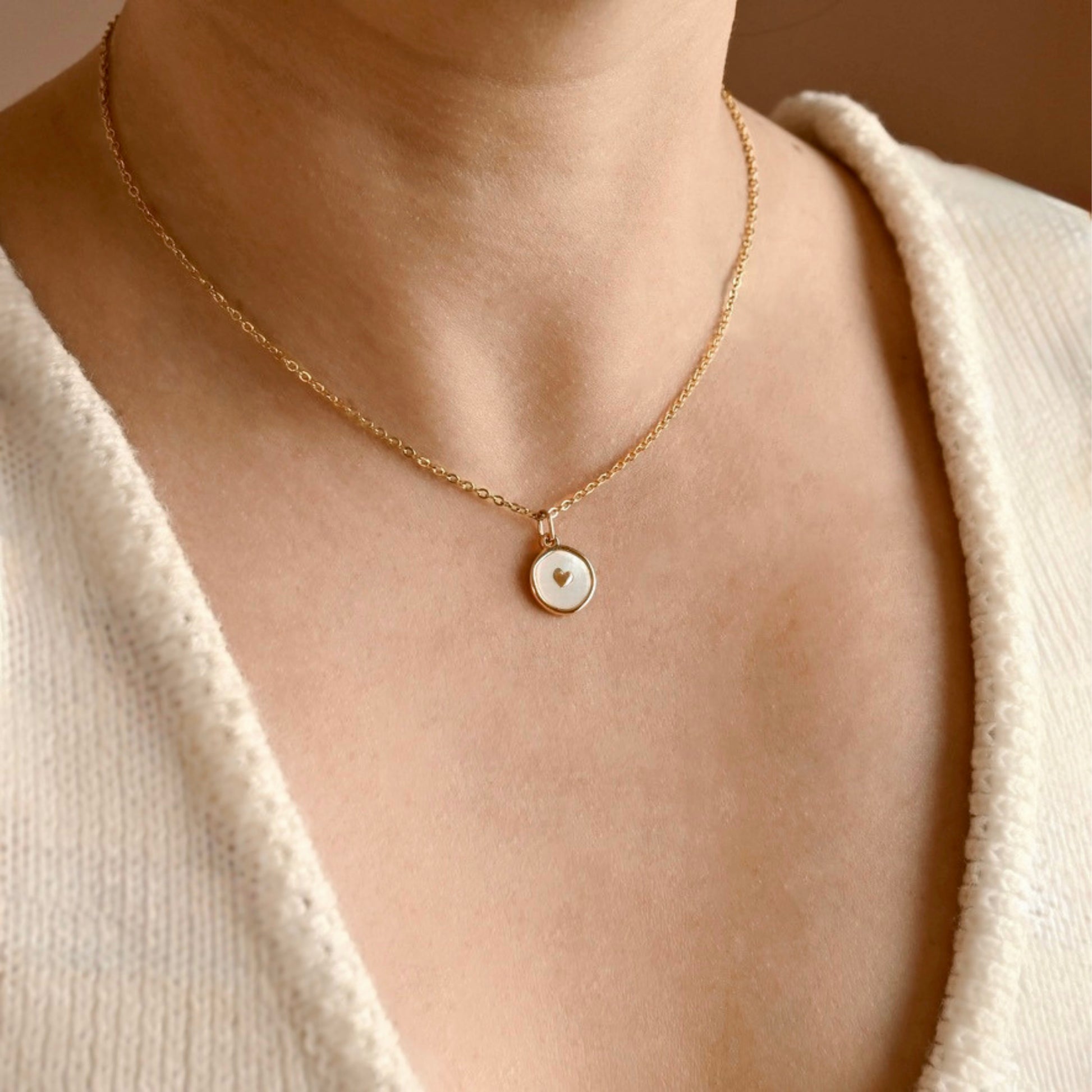 A close-up of a person's neck and upper chest wearing a delicate gold chain necklace with a small round pendant featuring a white inlay and a tiny gold heart.