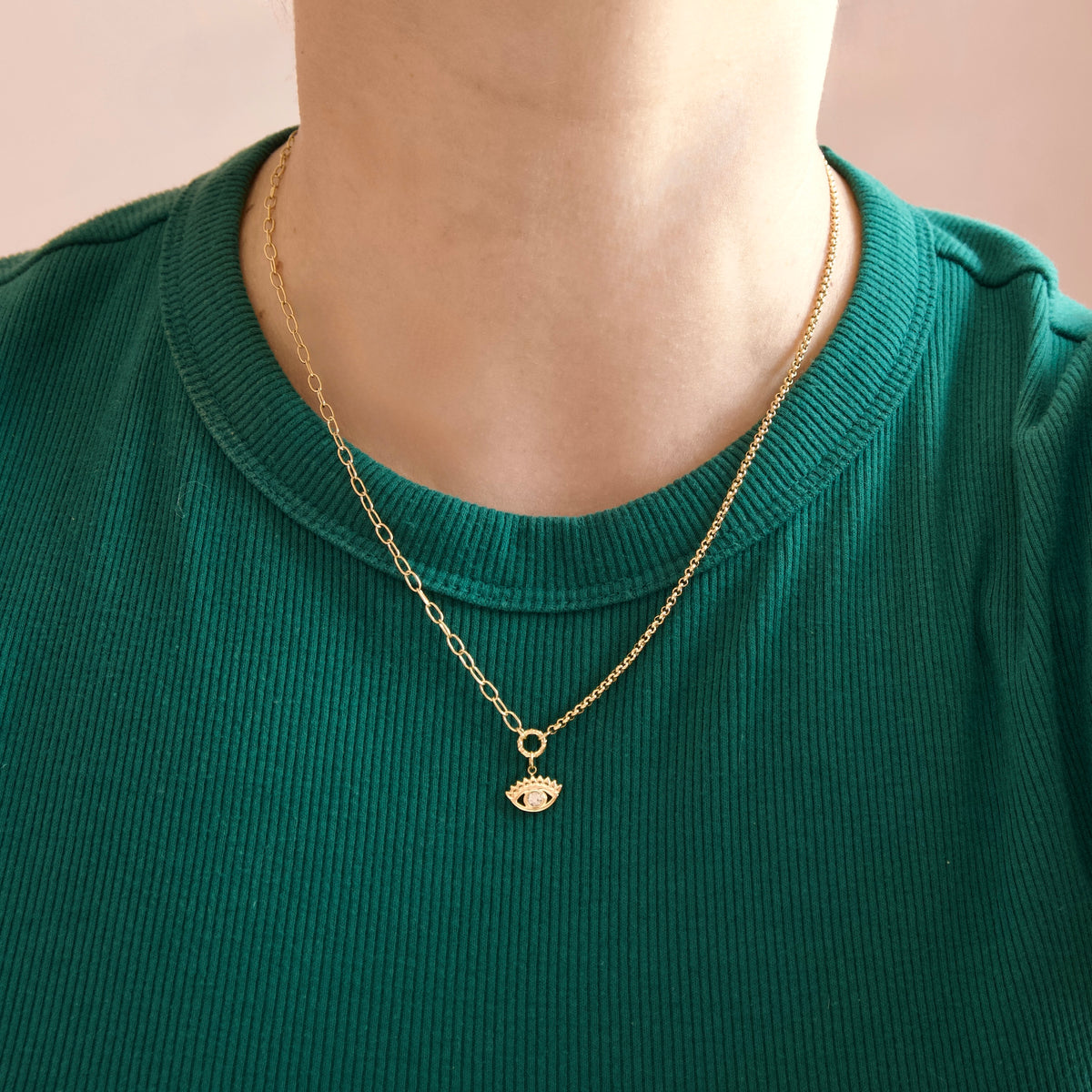 A close-up of a person wearing a delicate gold chain necklace with a small eye-shaped pendant, styled over a green ribbed t-shirt.