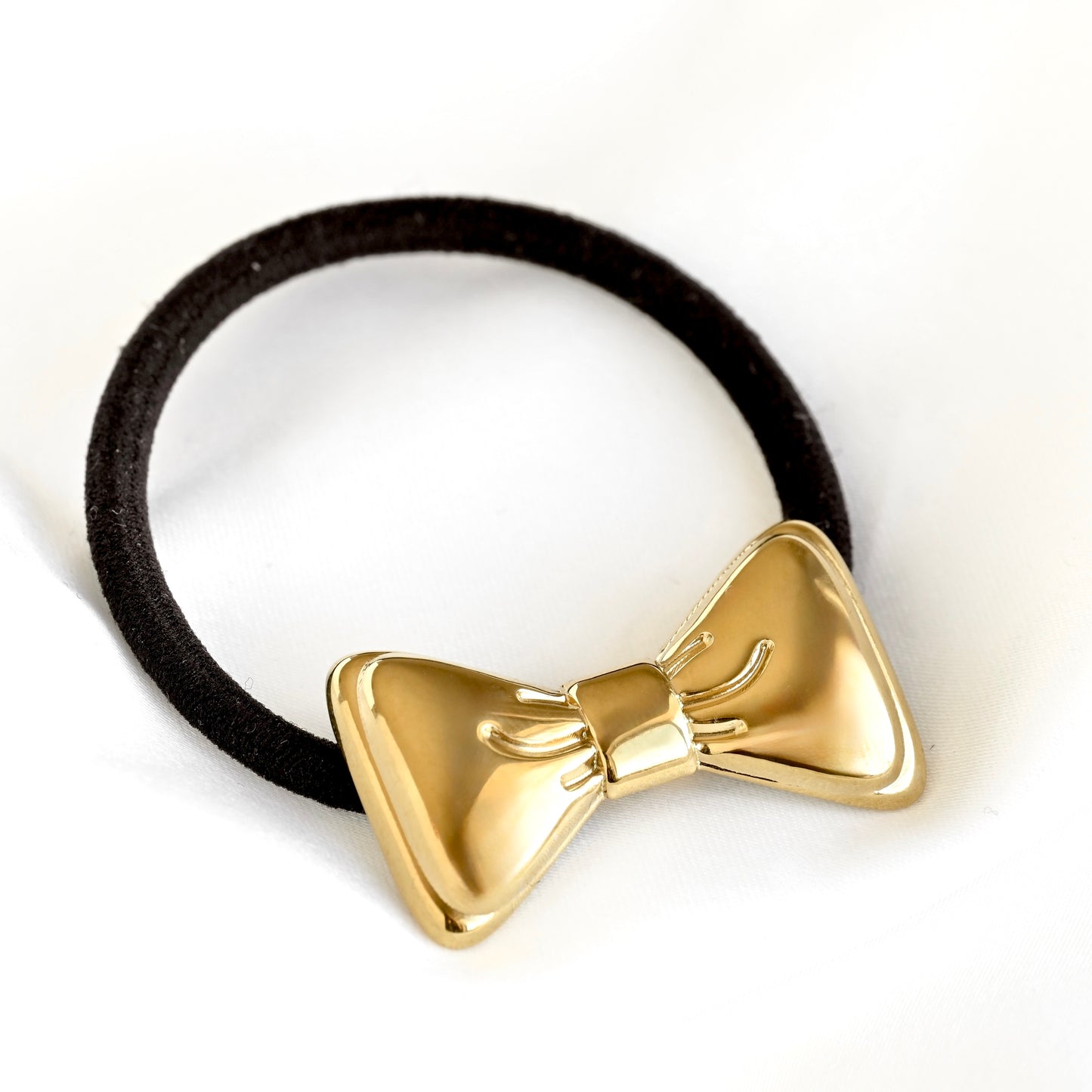 A black elastic hair tie with a shiny gold bow-shaped metal ornament, shown on a plain white background.