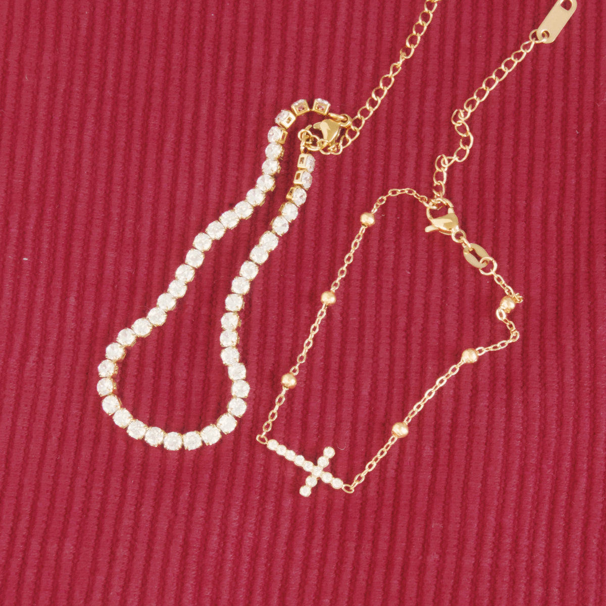 A gold chain bracelet with a small CZ cross charm and a tennis bracelet, displayed on a textured dark red ribbed fabric background.