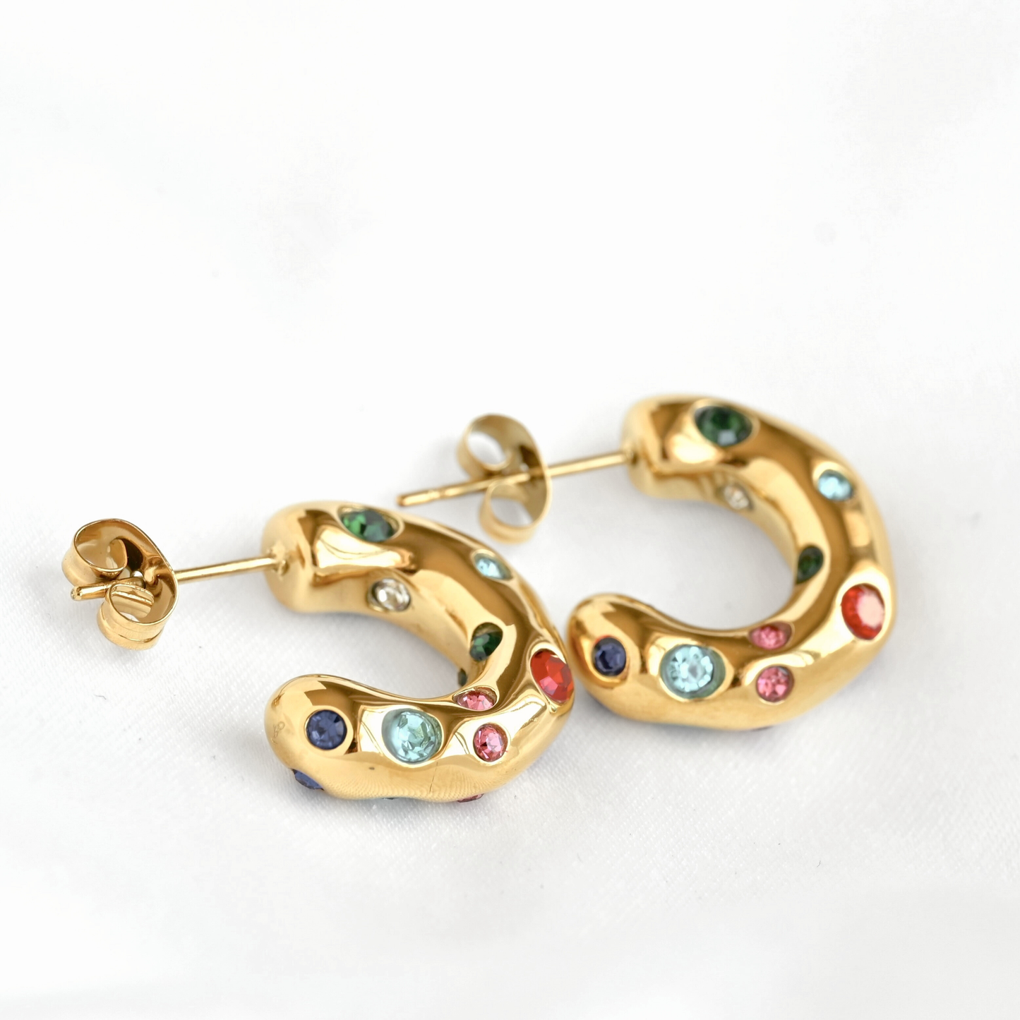 Gold hoop earrings with curved organic shapes, decorated with multiple small colorful gemstones in green, blue, pink, red, and dark blue, shown on a white background.